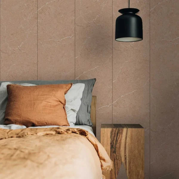 A styled bedroom corner featuring a bed with a rust-colored pillow and beige throw blanket, set against a wall of large vertical taupe stone tiles. A black pendant light hangs above, and a rustic wooden side table sits beside the bed.