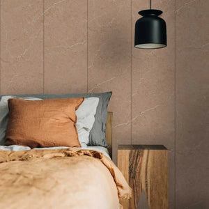 A styled bedroom corner featuring a bed with a rust-colored pillow and beige throw blanket, set against a wall of large vertical taupe stone tiles. A black pendant light hangs above, and a rustic wooden side table sits beside the bed.