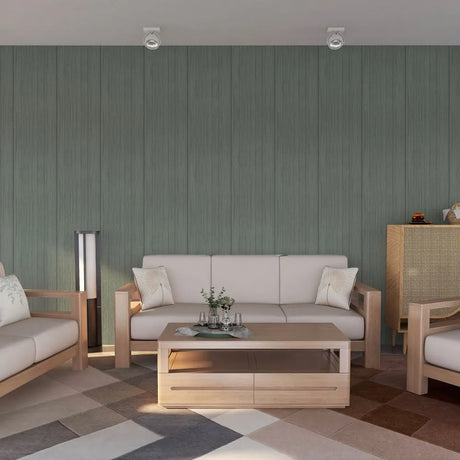 A modern living room setup featuring a light wood three-seater sofa with off-white cushions, a matching low wooden coffee table, and a geometric patterned rug. The background is a prominent sage green wood-paneled wall.
