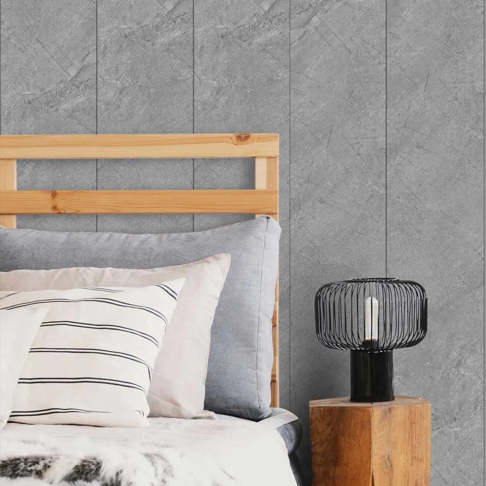 Gray textured wall paneling installed on a bedroom wall behind a wooden bed headboard, pillows, and a bedside table with a black wire lamp.