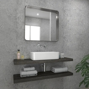 A modern bathroom vanity setup with a rectangular white vessel sink, chrome faucet, a large rounded-corner square mirror, and two dark floating shelves holding folded towels and toiletries against a textured gray wall.