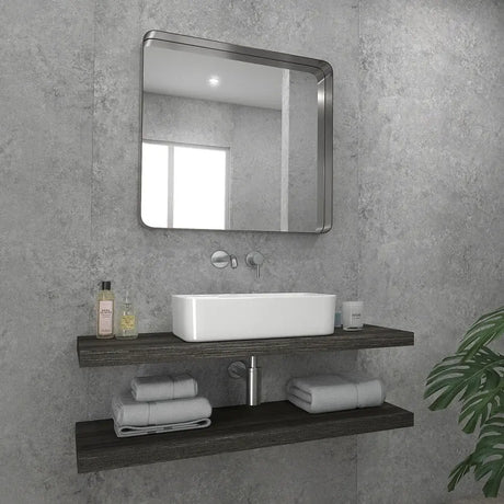 A modern bathroom vanity setup with a rectangular white vessel sink, chrome faucet, a large rounded-corner square mirror, and two dark floating shelves holding folded towels and toiletries against a textured gray wall.